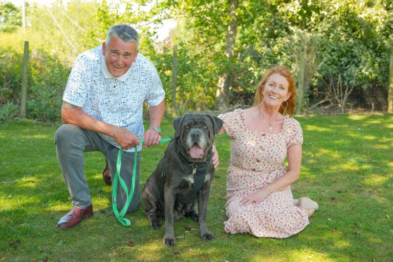 A couple kneel on a grass lawn with their large dog.