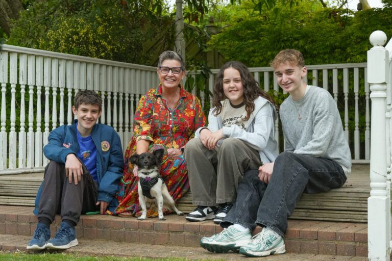 A family of four - a mum, daughter and two sons sit on a wooden decking with their small white and black dog. The full family looks directly at the camera, smiling.