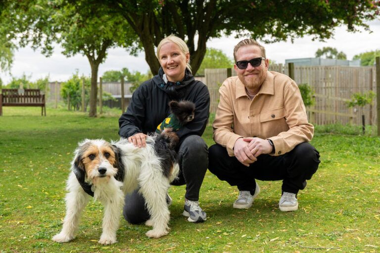 A blonde woman in a dark outfit holds a small black dog and sits next to a man wearing sunglasses, a tan jacket and dark jeans, a white, brown and black terrier dog sits in front of them. The whole group are sitting on a green lawn with trees in the background.